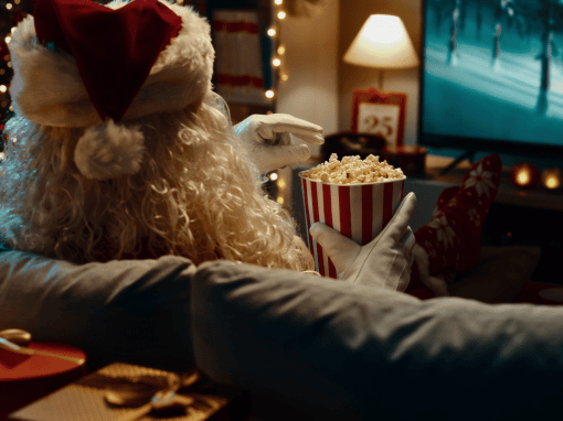 Santa Claus relaxing on a couch, eating popcorn while watching TV in a softly lit room decorated with Christmas lights.