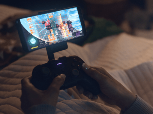 A close-up of hands holding a Luna Controller connected to an Android phone via the phone clip accessory, showing a game in progress in a softly lit bedroom at night.