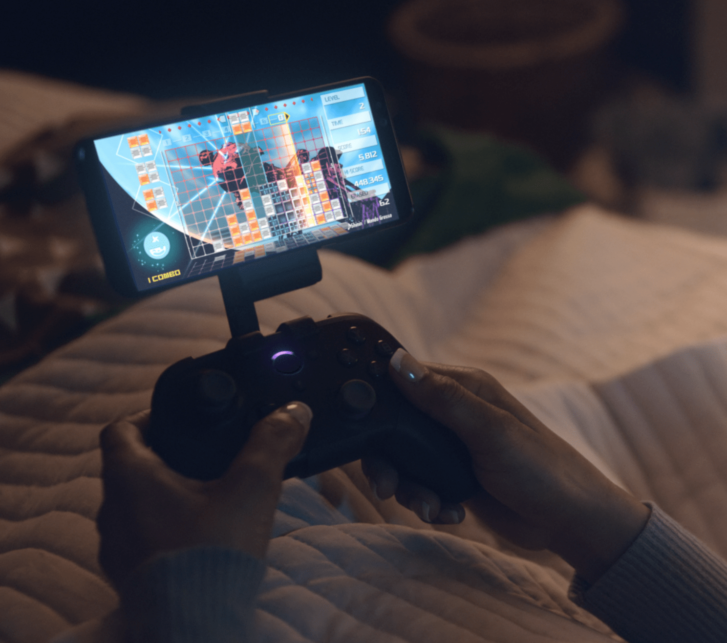 A close-up of hands holding a Luna Controller connected to an Android phone via the phone clip accessory, showing a game in progress in a softly lit bedroom at night.