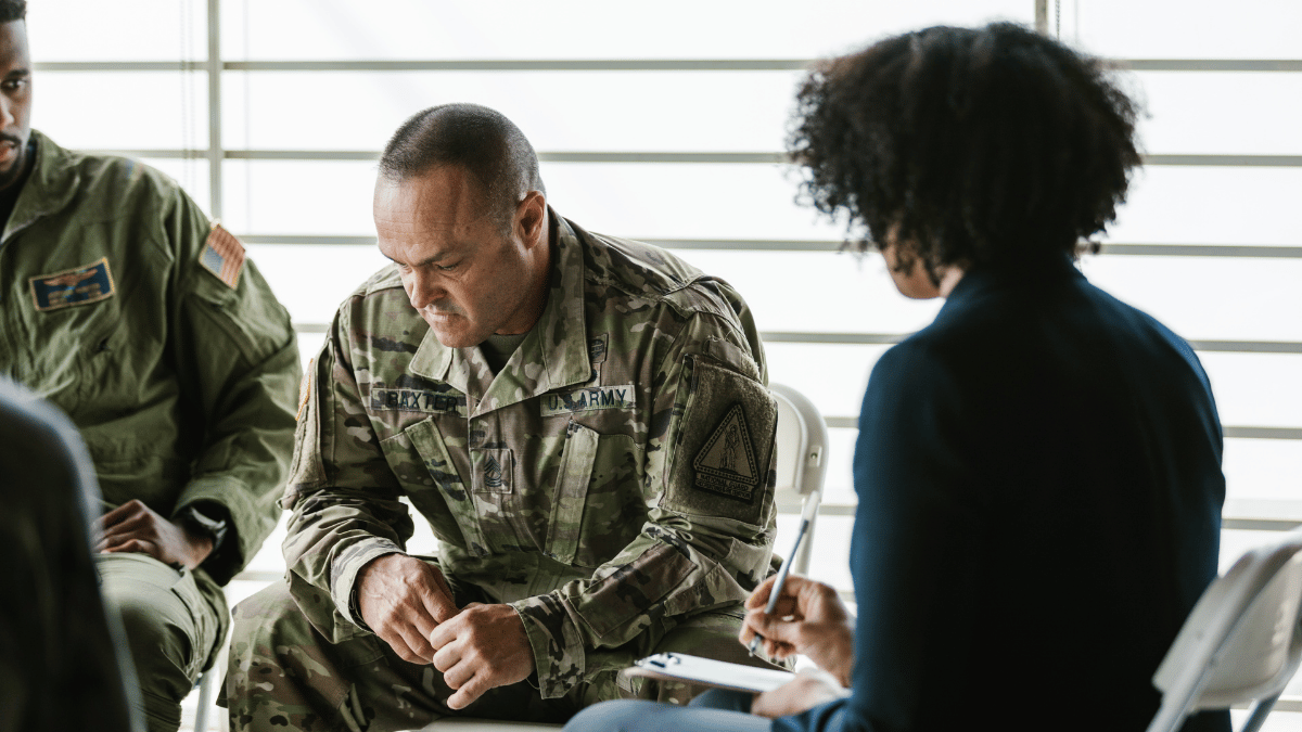 A veteran sits slumped in deep thought while a counselor and fellow veterans offer support, reflecting the emotional struggles many face after returning home.