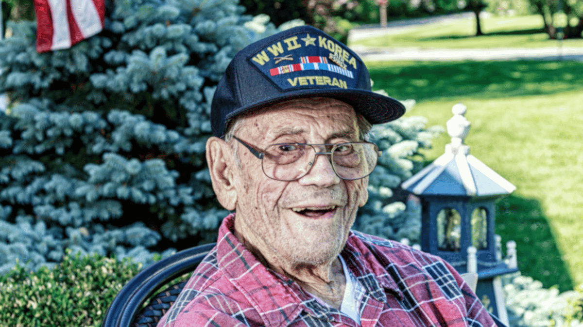 An elderly veteran wearing a ball cap showing service in both WWII and the Korean War, highlighting how Korea is often overlooked despite his sacrifice.