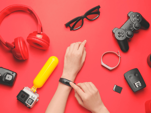 Assorted refurbished tech devices including headphones, camera, game controller, and a smartwatch being tapped by a woman’s finger on a pink tabletop.