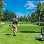 Golfer standing on the green holding a driver and looking down a sunny fairway, with golf bag and clubs beside him on a scenic course
